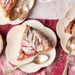 Three slices of Air Fryer Rhubarb Cake with powdered sugar rest on white plates, each with a spoon, set atop a red cloth and accompanied by a small bowl of extra powdered sugar.