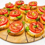 Slices of toasted bread topped with mozzarella, basil leaves, tomato slices, and a drizzle of balsamic glaze make this Caprese Appetizer irresistible on a wooden serving board.