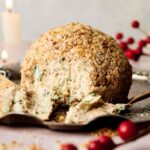 A Cheese Ball coated in chopped nuts sits on a decorative platter with a portion scooped out; crackers and red berries add a festive touch in the background.