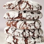 A stack of four Chocolate Crinkle Cookies, dusted with powdered sugar, sits temptingly on a white surface.