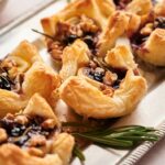 Golden puff pastry appetizers, like Cranberry Brie Bites, are topped with dark preserves, walnuts, and herbs on a silver tray, garnished elegantly with a sprig of rosemary.