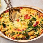 Baked vegetable frittata in a white oval dish with a serving spoon and a slice removed, revealing peas, spinach, tomatoes—an inviting twist on an Egg and Potato Breakfast Casserole.