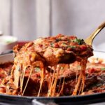 A serving spoon lifts a portion of cheesy baked Eggplant Casserole from a casserole dish, with melted cheese stretching from the pan.