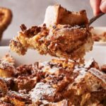 A close-up of French Toast Casserole being served from a baking dish, dusted with powdered sugar.