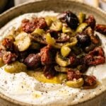 A bowl of whipped feta and goat cheese dip topped with chopped green and black olives, sun-dried tomatoes, olive oil, and black pepper.