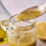 A spoon hovers over a glass jar filled with Lemon Vinaigrette Dressing; a halved lemon and a bottle of oil are visible in the background.