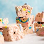Several pink Lucky Charm Rice Krispy Treats with colorful candy pieces are stacked on a white surface with a light blue background.