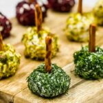 An assortment of Mini Goat Cheese Balls coated with herbs, nuts, and seeds, each with a pretzel stick inserted, arranged elegantly on a wooden board.