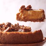 A slice of Pecan Pie Cheesecake topped with glazed pecans is being lifted from a whole cheesecake on a plate.