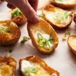 A hand lifts a Loaded Potato Skin filled with melted cheese, sour cream, and chopped green onions from a tray packed with similar delicious potato skins.
