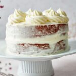 A partially frosted Red Velvet Cake with white buttercream icing, decorated with swirls of frosting on top, sits elegantly on a white cake stand.