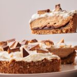 A slice of Reese's Peanut Butter Pie with a crumbly crust, creamy filling, whipped topping, and chocolate pieces is being lifted from the whole pie on a plate.