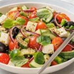 A bowl of Southern Farmhouse Feta & Veggie Salad features feta cheese, cherry tomatoes, cucumber, black olives, red onion, and fresh mint leaves, served with a fork.