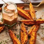 Sweet Potato Wedges oven-baked to perfection rest on parchment paper, accompanied by a jar of creamy dipping sauce with a spoon.