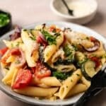 Plate of vegetable pasta with penne and roasted vegetables, topped with grated cheese and chopped herbs. A fork rests on the side, with small bowls of extra cheese and herbs in the background.