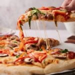 A hand lifts a slice of Veggie Pizza with melted cheese, mushrooms, red onions, and bell peppers from the whole pizza on a table.