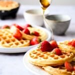 Plates of golden waffles topped with fresh strawberries, with honey being drizzled from a spoon; cups and a waffle maker in the background.