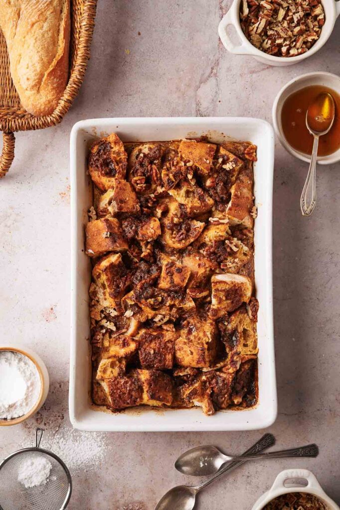 A rectangular baking dish filled with French Toast Casserole sits on a countertop, surrounded by a loaf of bread, bowls of pecans, syrup, and powdered sugar, with two spoons nearby.