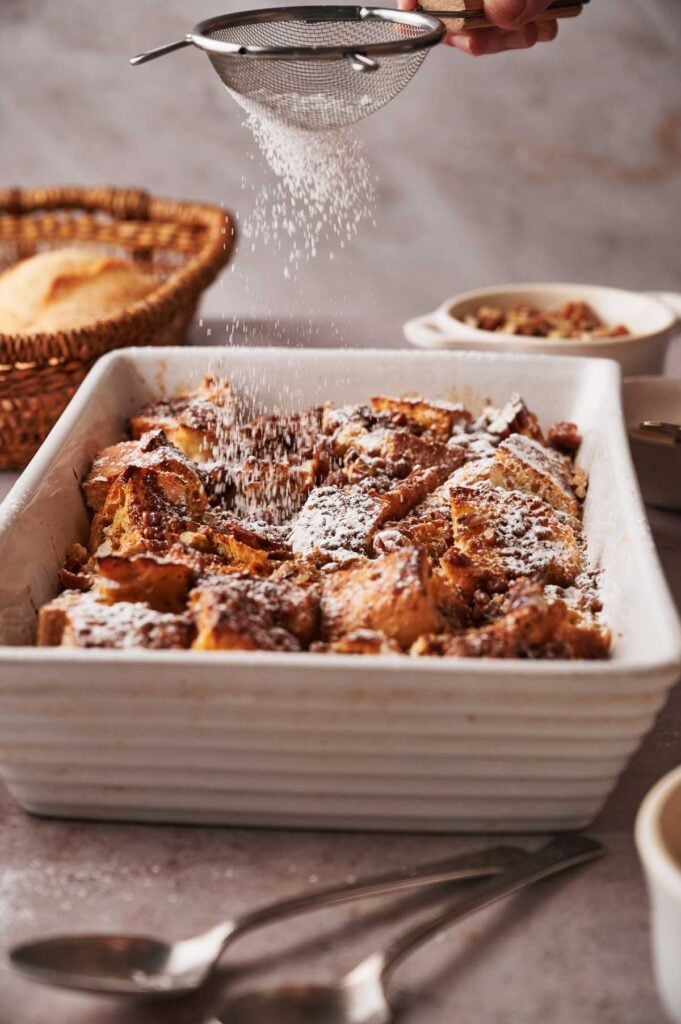 A hand sifts powdered sugar over a freshly baked french toast casserole in a white dish, with bread and utensils visible in the background.