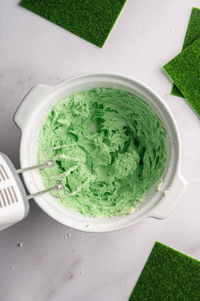 A white mixing bowl with green frosting being mixed by an electric hand mixer, surrounded by green square pieces on a white surface—perfect for decorating Football Cupcakes.
