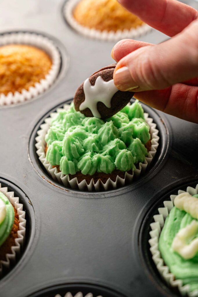 A hand places an iced chocolate cookie on a Football Cupcake with green frosting in a muffin tin, surrounded by other decorated cupcakes.