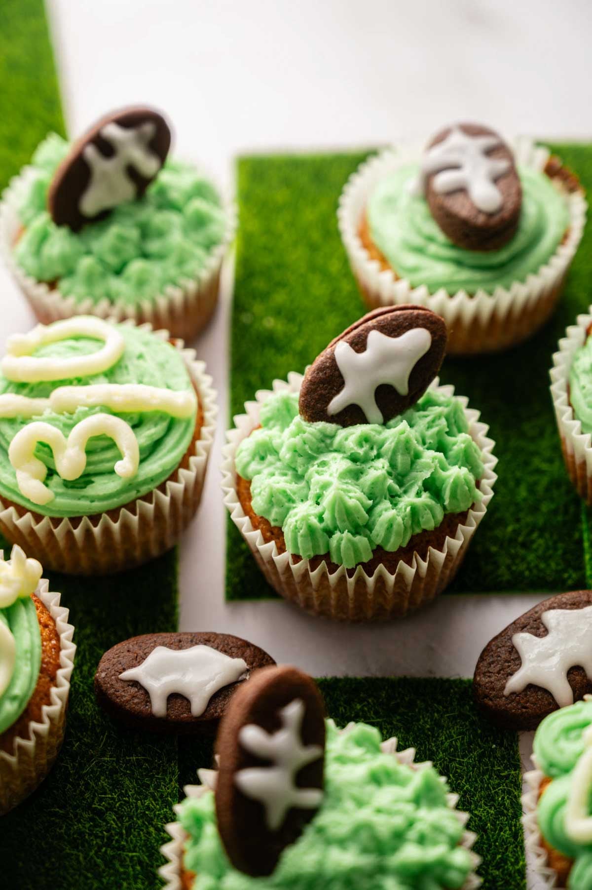 Football Cupcakes with green frosting are decorated with football-shaped cookies and piped white icing, all displayed on a surface resembling artificial grass.