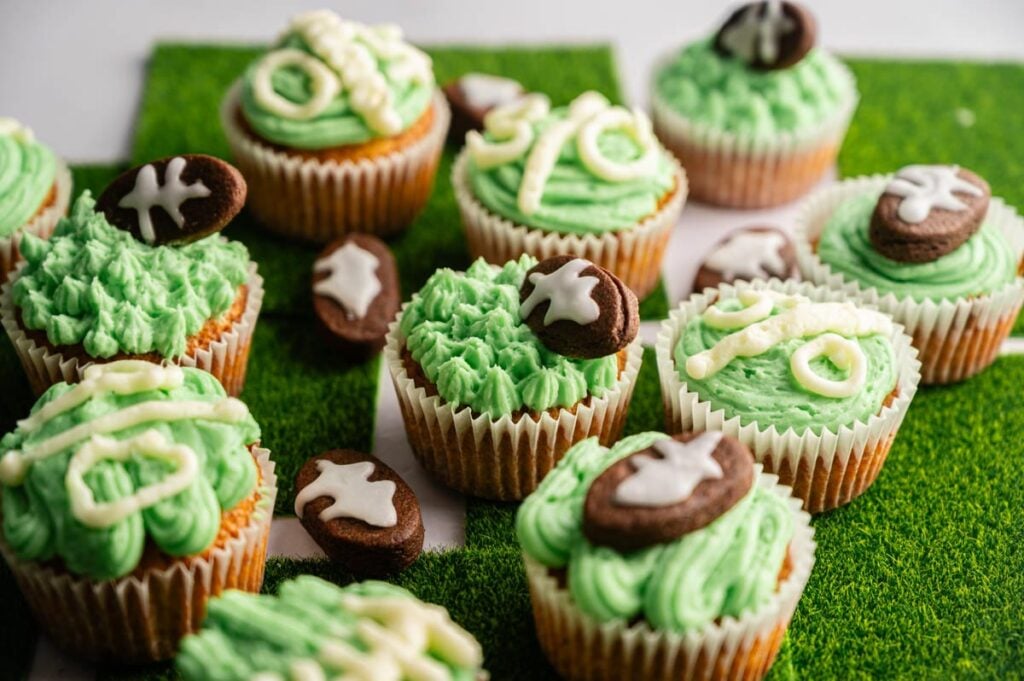 Football Cupcakes with green frosting and football-themed decorations are arranged on a faux grass surface. Some cupcakes feature icing shaped like footballs or game play diagrams, perfect for game day celebrations.