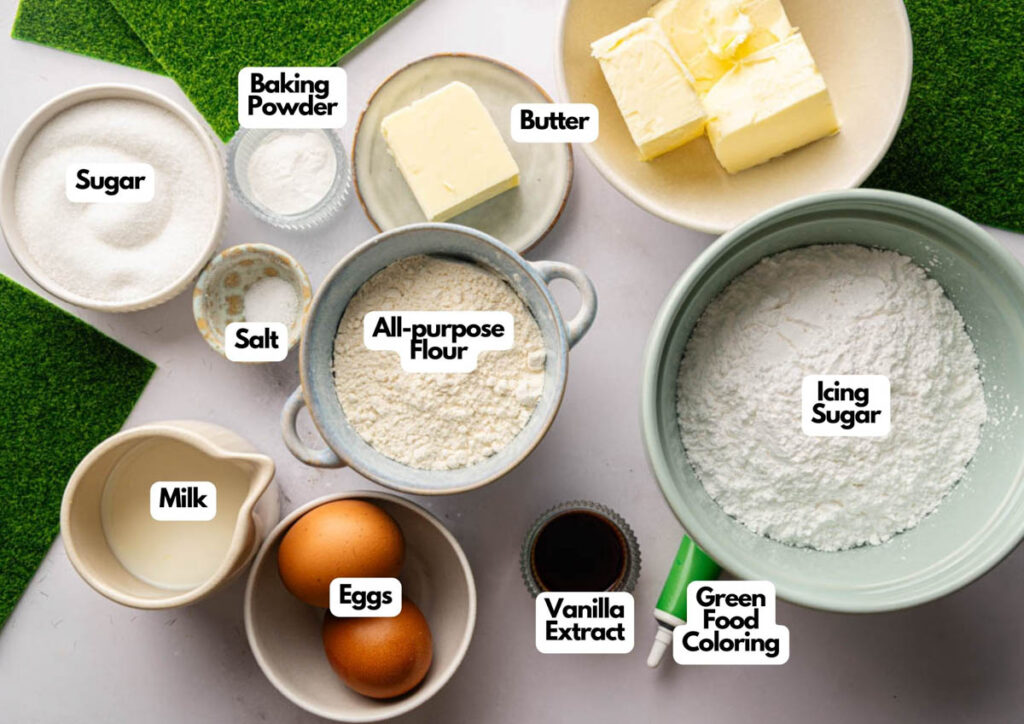Various baking ingredients for Football Cupcakes—including sugar, salt, milk, eggs, baking powder, all-purpose flour, butter, vanilla extract, icing sugar, and green food coloring—are neatly arranged on a countertop.