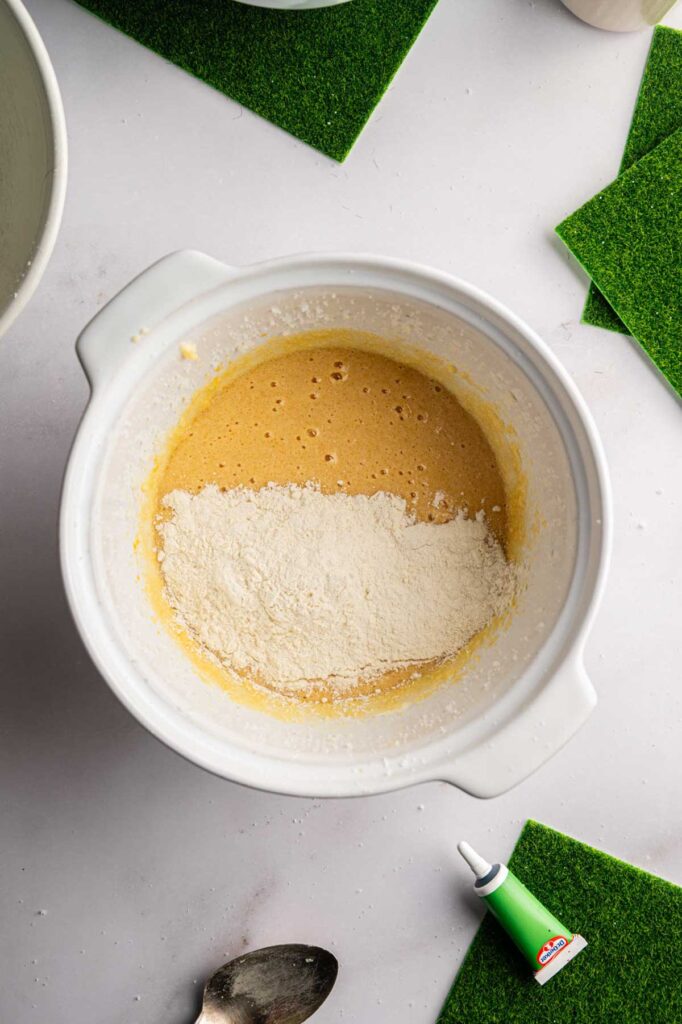 A white mixing bowl contains a yellow batter with a layer of flour on top, ready to be transformed into festive Football Cupcakes. A spoon and a green food coloring tube are nearby on a white surface with green felt squares.