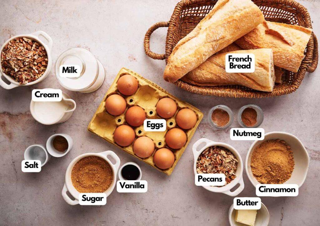 Top-down view of ingredients labeled for a French Toast Casserole recipe, featuring French bread, eggs, milk, cream, salt, sugar, vanilla, pecans, nutmeg, cinnamon, and butter arranged on a countertop.