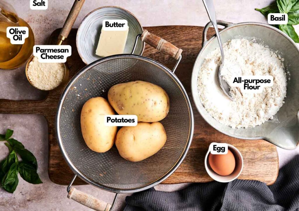 Labeled ingredients for making Air Fryer Gnocchi, including potatoes, flour, egg, butter, parmesan cheese, olive oil, salt, and fresh basil, arranged on a kitchen surface.