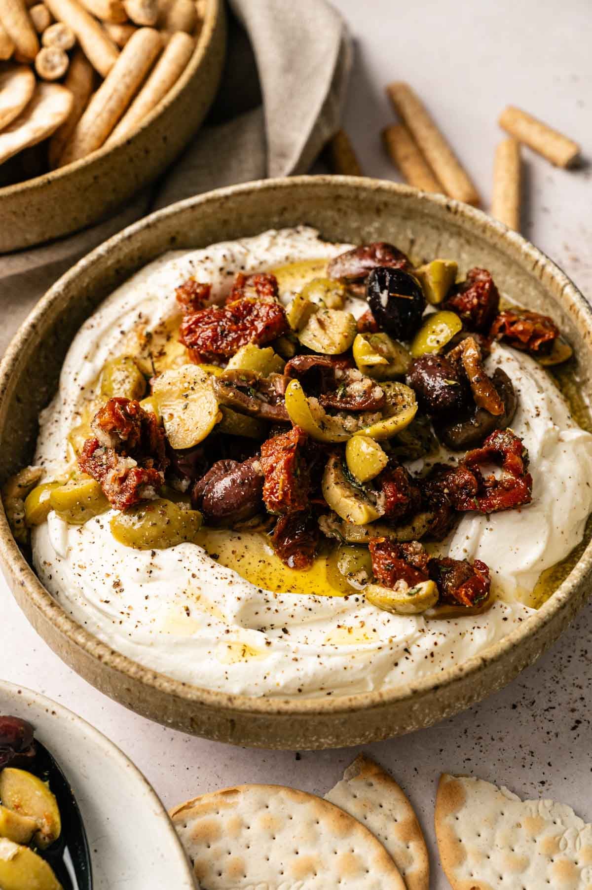 A bowl of creamy Goat Cheese Dip topped with sliced olives, sun-dried tomatoes, and olive oil, surrounded by crackers and breadsticks.