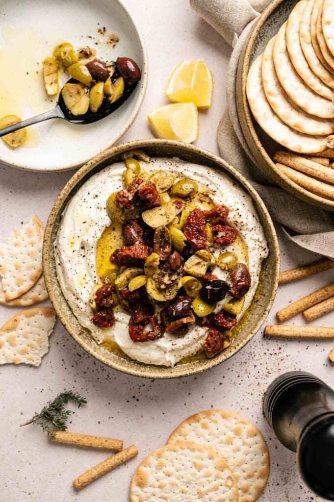 A bowl of whipped feta and creamy goat cheese dip topped with chopped olives and sun-dried tomatoes, surrounded by crackers, breadsticks, and lemon wedges on a light surface.