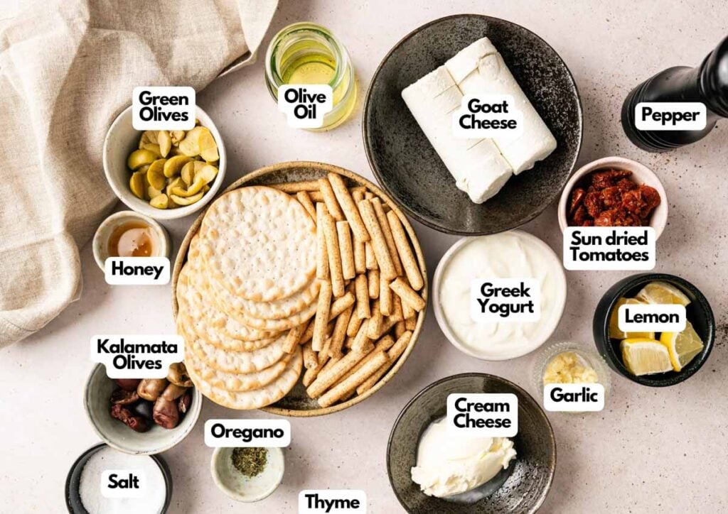 Assorted Mediterranean ingredients and snacks including Goat Cheese Dip, Greek yogurt, cream cheese, olives, crackers, herbs, honey, sun-dried tomatoes, lemon, salt, garlic, and pepper.