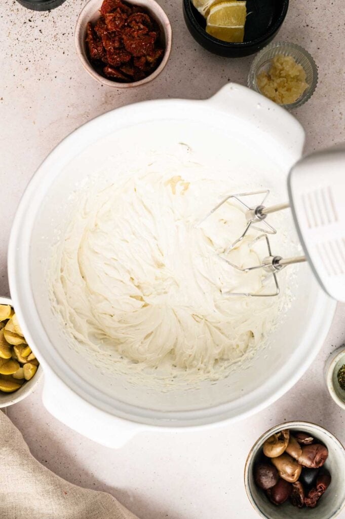 Electric hand mixer in a white bowl filled with whipped creamy mixture for Goat Cheese Dip, surrounded by small bowls of various ingredients on a light countertop.