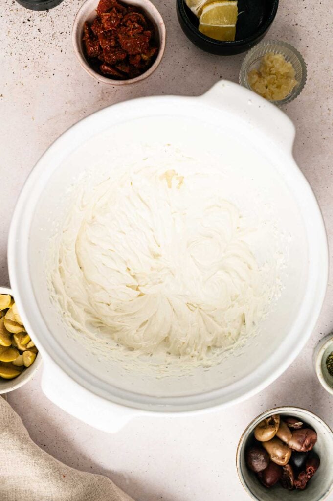 A mixing bowl with whipped goat cheese dip in the center, surrounded by small bowls of sun-dried tomatoes, lemon, garlic, olives, and sliced pickles.