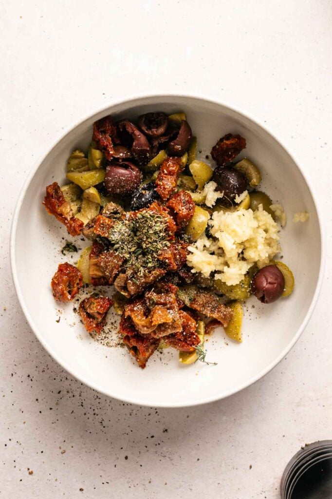 A white bowl contains chopped green and black olives, minced garlic, sun-dried tomatoes, and dried herbs, all sprinkled with black pepper—the perfect Mediterranean mix to complement a creamy Goat Cheese Dip.