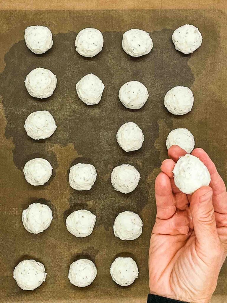 A hand holds a ball of dough above a baking sheet lined with rows of evenly spaced goat cheese balls, all ready for baking.