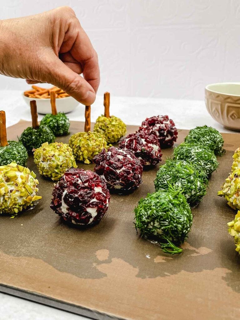 A hand places a goat cheese ball coated in chopped pistachios among other goat cheese balls coated with herbs and cranberries, all with pretzel sticks inserted.