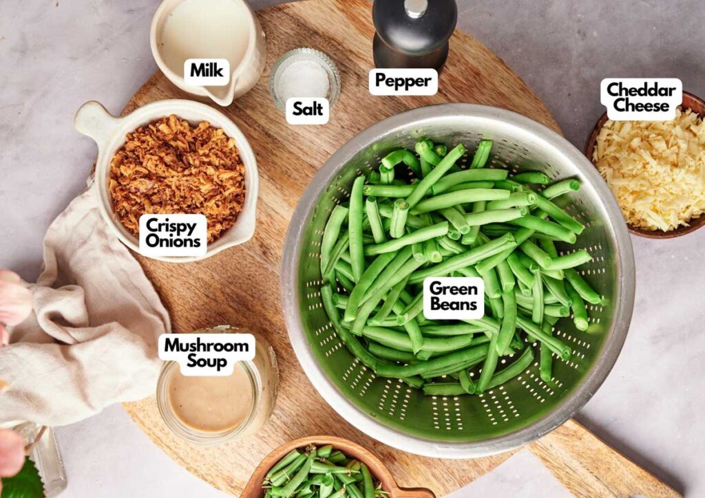 A bowl of green beans in a colander is surrounded by bowls of milk, cheddar cheese, crispy onions, mushroom soup, salt, and a pepper grinder&mdash;everything you need to create a classic Green Bean Casserole.