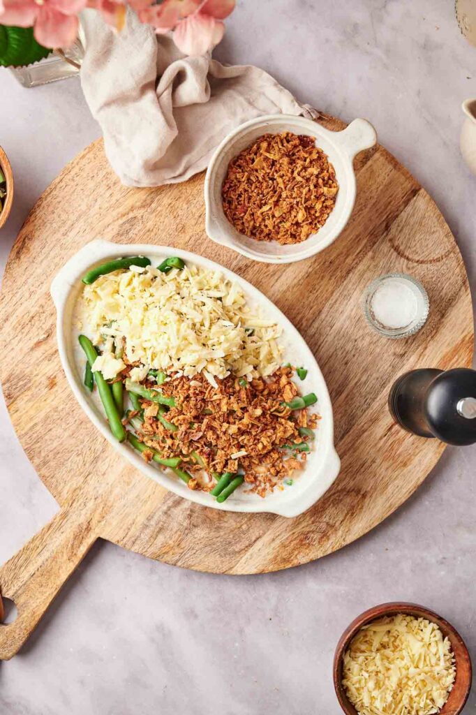 Oval dish of Green Bean Casserole topped with shredded cheese and crispy onions, served on a wooden board beside a bowl of extra crispy onions, a napkin, salt, and a pepper grinder.