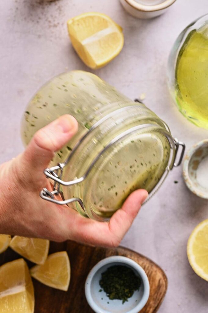 A hand holds a glass jar with a clasp lid containing a green herb mixture, perfect for Lemon Vinaigrette Dressing; lemon wedges and small bowls are nearby.