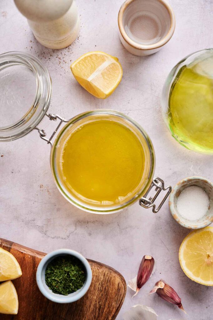 Top-down view of a jar filled with yellow Lemon Vinaigrette Dressing, surrounded by lemon wedges, herbs, garlic, a salt dish, oil, and a pepper grinder on a light countertop.
