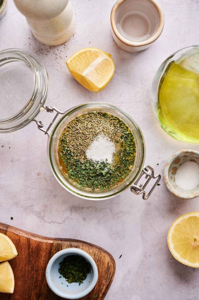 Dried thyme, basil, and salt sit on top of the lemon and olive oil base in a jar, ready to be shaken together.