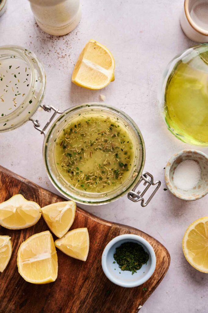 Glass jar of Lemon Vinaigrette Dressing on a white surface, surrounded by lemon wedges, a bowl of dried herbs, salt, and a bottle of olive oil.