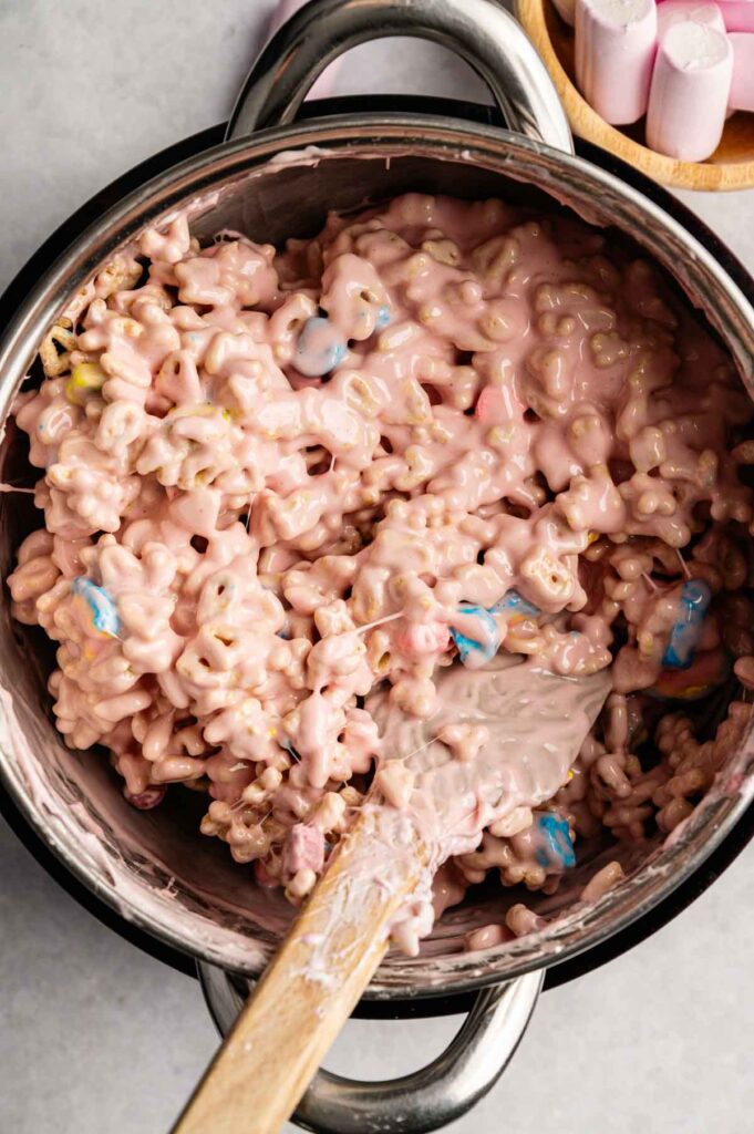 A pot filled with pink, gooey Lucky Charm Rice Krispy Treat mixture, with visible pieces of cereal and pastel-colored marshmallows, and a spatula resting inside.