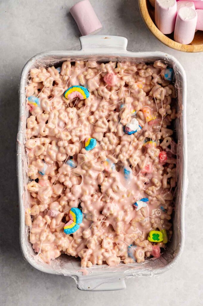 A baking dish filled with Lucky Charm Rice Krispy Treats, topped with colorful marshmallow shapes; a bowl and loose pink marshmallows are beside the dish.