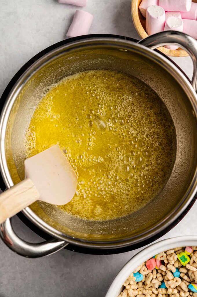 A saucepan with melted butter being stirred by a spatula, surrounded by bowls of marshmallows and colorful cereal on a gray surface, ready for making delicious Lucky Charm Rice Krispy Treats.