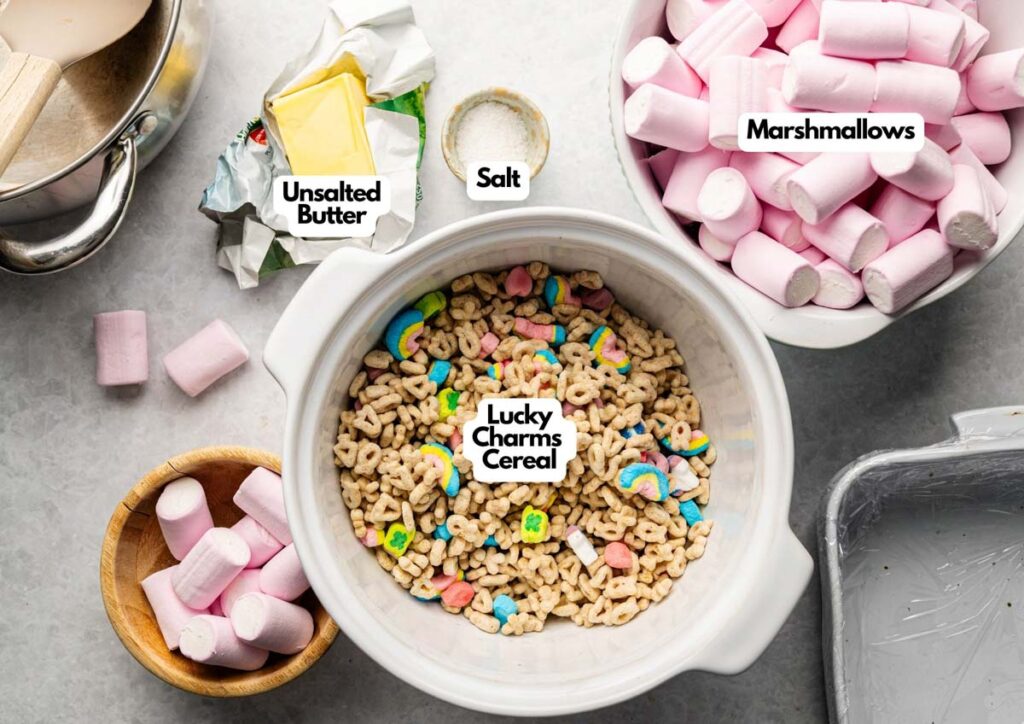 A bowl of Lucky Charms cereal, a dish of unsalted butter, pink and white marshmallows, a small bowl of salt, and a pot are arranged on a gray surface&mdash;perfect for making homemade Lucky Charm Rice Krispy Treats.