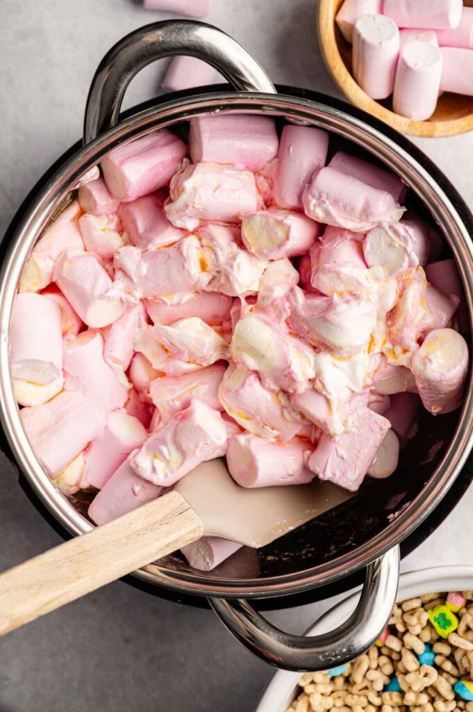A pot filled with marshmallows being stirred with a spatula, perfect for making Lucky Charm Rice Krispy Treats, with a bowl of cereal and more marshmallows nearby.
